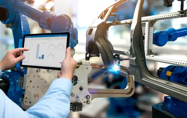 Ein Roboter schweißt ein Autoteil, während ein Mensch auf einem Tablet die Produktionsdaten prüft. Was ist eine BOM? Hier wird sie digital verwaltet.